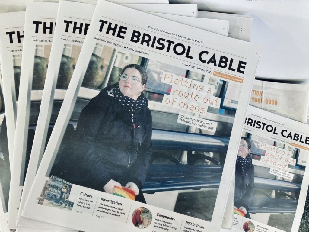 The front cover of Edition 32 of The Bristol Cable. A woman is sitting at a bus stop, waiting. The headline is 'Plotting a route out of chaos: Could franchising end Bristol's bus nightmare?'