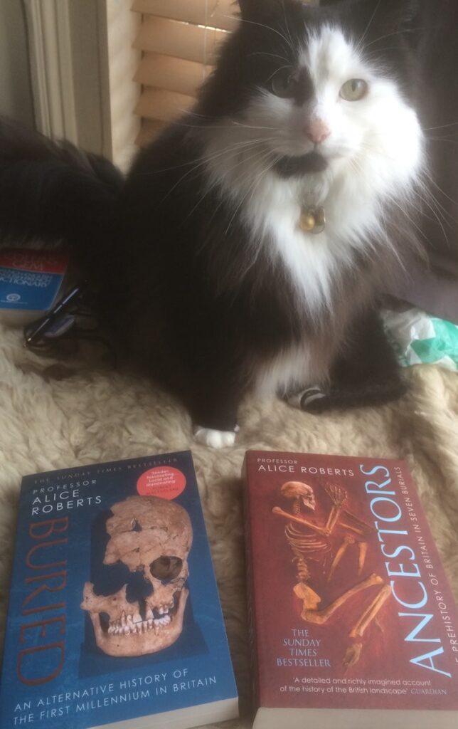 Black & white cat standing behind the books Buried and Ancestors by Professor Alice Roberts.