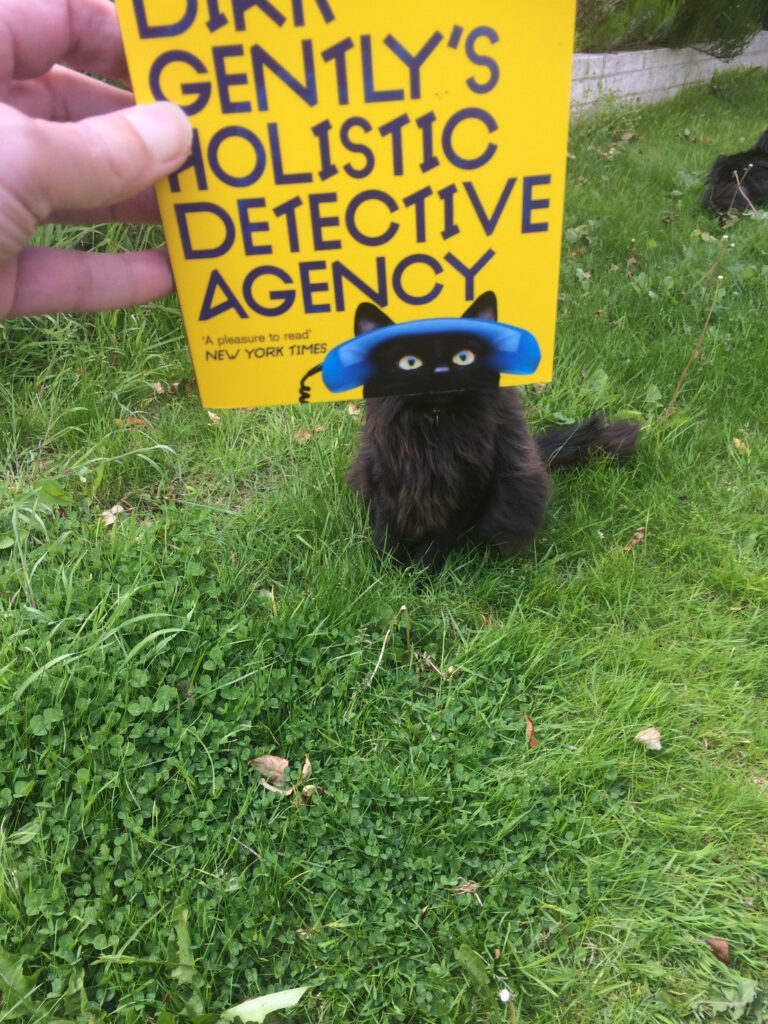 A copy of Dirk Gently's Holistic Detective Agency, held out so that the cut-off image at the bottom of the cover, of the top half of a black cat's head with a blue telephone receiver perched across it, aligns with a black cat sat on a lawn behind the book.
