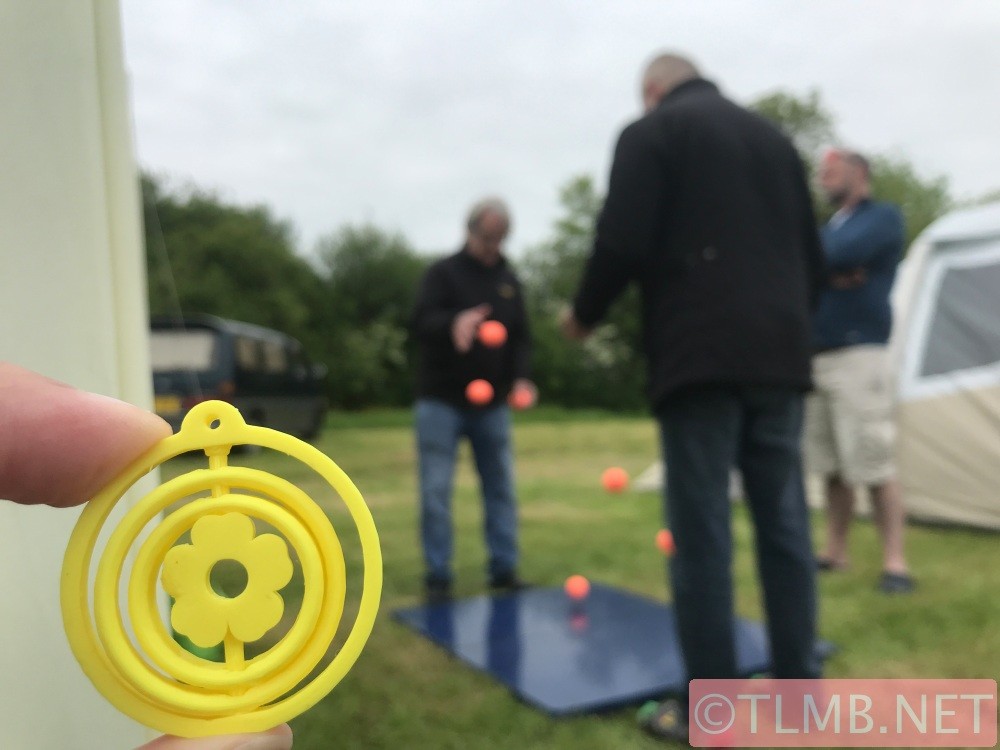 A yellow plastic buttercup within 3 spinnable rings. In the background two jugglers are bounce-passing a bunch of orange balls on a marble slab in the middle of a field.